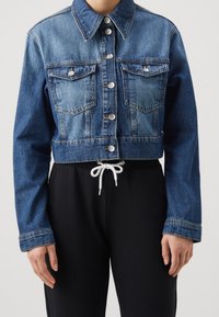 Krátká džínová bunda ve středně modré barvě, s zapínáním na knoflíky, dvěma náprsními kapsami, klasickým límečkem a kontrastními černými tepláky.