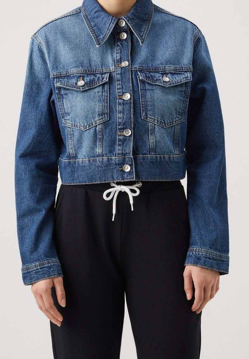 Krátká džínová bunda ve středně modré barvě, s zapínáním na knoflíky, dvěma náprsními kapsami, klasickým límečkem a kontrastními černými tepláky.