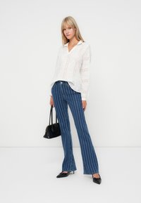 White, eyelet blouse with a collar, paired with blue flared jeans featuring vertical white stripes. Model holds a small black handbag.
