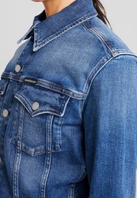 Detailní záběr na modrou džínovou bundu s kovovými knoflíky s logem Calvin Klein a malou nášivkou Calvin Klein Jeans na kapsičce na hrudi.