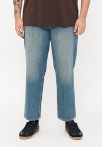 Heledad helesin denim sirge jalgadega teksad helesinises toonis, millel on fadeeritud muster, töötlemata serv ja kulunud välimus, koos mustade sportkingadega.