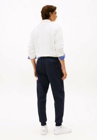 Weißes Sweatshirt mit aufgerollten Ärmeln, blau gestreiften Bündchen, marinefarbene Jogginghosen mit rot-weißen Akzenten und weißen Sneakers. Rückansicht des Outfits.