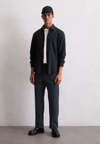 Man wearing black cap, black button-up shirt over white tee, dark pants, and black loafers standing against a light textured wall.