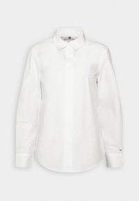 Chemise blanche à manches longues boutonnée avec col, petit écusson brodé sur la poitrine gauche et logo sur la manchette droite, présentée sur un fond uni.
