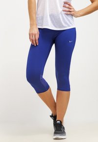 Kvinna som bär blå Nike knälånga leggings, vit topp och svarta Nike löparskor, står med ena handen på höfterna och korsade ben.