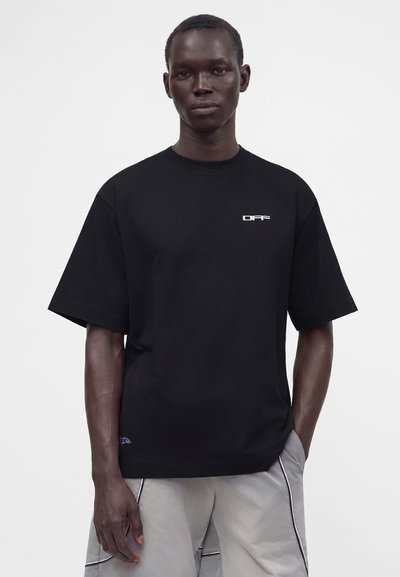 Άνδρας που φοράει μαύρο φαρδύ t-shirt με το λογότυπο "OFF" στο στήθος και ανοιχτόχρωμο γκρι σορτς με μαύρη επένδυση, στέκεται μπροστά σε απλή φόντο.