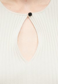 Κοντινό πλάνο ενός λευκού ριμπ πλεκτού τοπ με άνοιγμα στο λαιμό σε σχήμα σταγόνας, ασφαλισμένο με ένα μικρό μαύρο κουμπί στον γιακά.