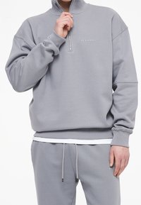 Sudadera de cremallera de color gris claro con cuello alto, puños y cinturilla de canalé; presenta el logotipo de la marca en un tono contrastante en el pecho.