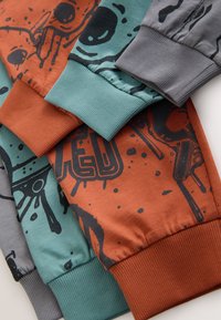 Trois paires de joggeurs de couleurs variées : bleu sarcelle avec des motifs noirs, orange rouille avec des lettres audacieuses, et gris avec des détails noirs. Chaque modèle est doté de poignets côtelés.