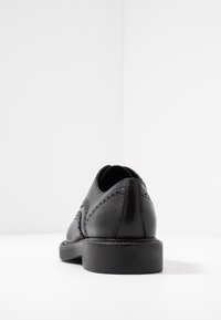 Chaussure en cuir noir à bout arrondi, présentant des coutures complexes et un dos texturé. Semelle plate en caoutchouc avec un détail de marque subtil.