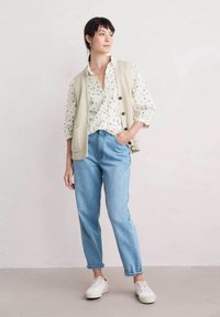 Stickad beige väst över en blommig skjorta med knappar, ihop med ljusblå jeans och vita sneakers, stående mot en neutral bakgrund.