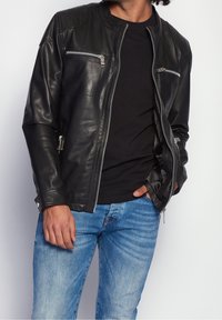 Giacca di pelle nera con una texture liscia, dotata di tasche sul petto con zip, chiusura con zip e dettagli a coste sulle spalle. Indossata sopra una camicia nera.