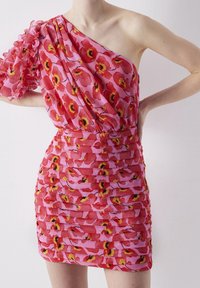 Femme portant une robe rose à une épaule avec un imprimé floral rouge et jaune et des détails à volants superposés, posant les mains sur les hanches.