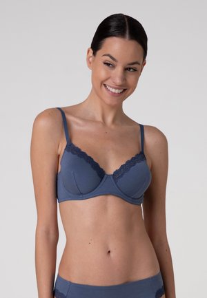 Giovane donna con capelli scuri in reggiseno blu con rifiniture in pizzo e biancheria intima coordinata, sorridente e che guarda di lato su sfondo neutro.