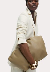 Borsa in pelle beige con un design morbido e imbottito. Portata sotto il braccio con un blazer bianco, caratterizzato da dettagli in metallo dorato sui polsini.
