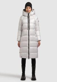 Világosszürke, bélelt hosszú kabát kapucnival, elülső cipzárral és fekete részletekkel. Fekete leggingszel és fűzős csizmával párosítva.