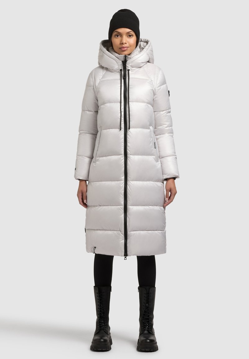 Világosszürke, bélelt hosszú kabát kapucnival, elülső cipzárral és fekete részletekkel. Fekete leggingszel és fűzős csizmával párosítva.