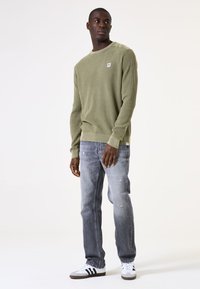 Olivgrüner Waffle-Knit-Pullover mit rundem Halsausschnitt und geripptem Saum, kombiniert mit grauen, verwaschenen Jeans und weißen Sneakern mit schwarzen Streifen.