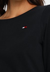 Schwarzes Langarmshirt aus weichem Stoff, mit einem kleinen gestickten Logo in Rot, Weiß und Blau auf der linken Seite.