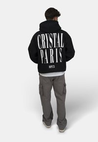 Svart huvtröja med stor vit text "CRYSTAL PARIS" på baksidan, kombinerad med grå cargobyxor. Huvtröjan har en dragsko och stor huva.