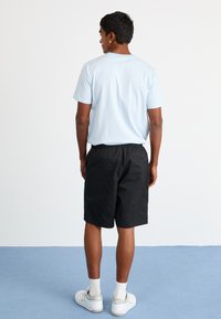 Lys blåstripet T-skjorte med kortermet; svarte shorts med elastisk midje; hvite sneakers med grå detaljer og hvite sokker; ensfarget bakgrunn.
