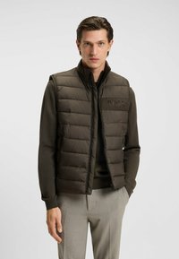 Braune gepolsterte Weste mit hohem Kragen, Reißverschluss und Logodetail. Kombiniert mit einem dunklen Pullover und hellgrauen Hosen.
