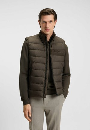 Gilet imbottito marrone con colletto alto, chiusura zip e dettaglio del logo. Indossato sopra un maglione scuro, abbinato a pantaloni grigio chiaro.