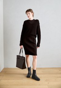 Pull en maille noire avec un col en V, jupe crayon noire, bottines chunky noires et un grand sac en toile sombre. Sol en bois et mur gris en arrière-plan.