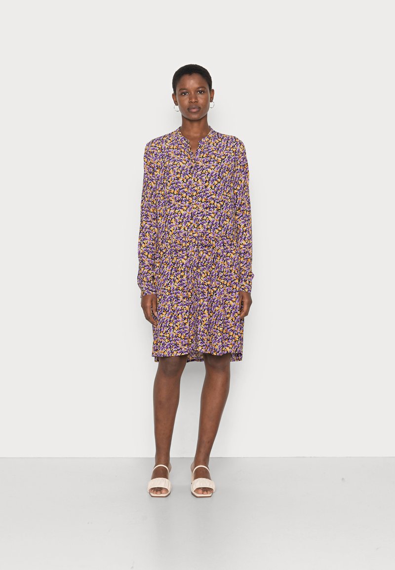 Vestido floral roxo com mangas longas, decote com botões e um ajuste solto, combinado com sandálias de cor clara.