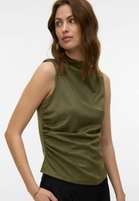 Femme aux cheveux bruns ondulés portant un top olive sans manches froncé et un pantalon noir, posant avec la main dans la poche devant un fond blanc.