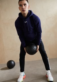 Marineblauwe hoodie met capuchon en voorzak, zwarte sportbroek met witte strepen en kleurrijke sneakers, terwijl hij een zwarte fitnessbal vasthoudt.