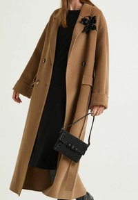 Cappotto in misto lana cammello con design a doppio petto, grandi polsini e un particolare fiore nero in paillettes. Abbinato a una piccola borsa nera con perline.