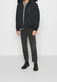 Sort puffersjakke med en pelsforet hætte, lynlås- og knaplukning, båret over en grå sweater og parret med mørke jeans og sorte ankelstøvler.