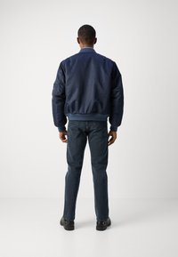 Marineblå bomberjakke med ribbede mansjetter og midjebånd, brukt over mørkeblå jeans, sett fra baksiden mot en enkel bakgrunn.