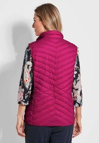 Taillierte, ärmellose Steppweste in lebhaftem Magenta mit einem Chevron-Steppmuster. Von hinten getragen über einer blumigen Langarmbluse.