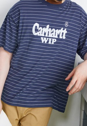 Homme portant un t-shirt rayé bleu marine avec le logo blanc "Carhartt WIP", associé à un pantalon beige, posant le bras sur un meuble.