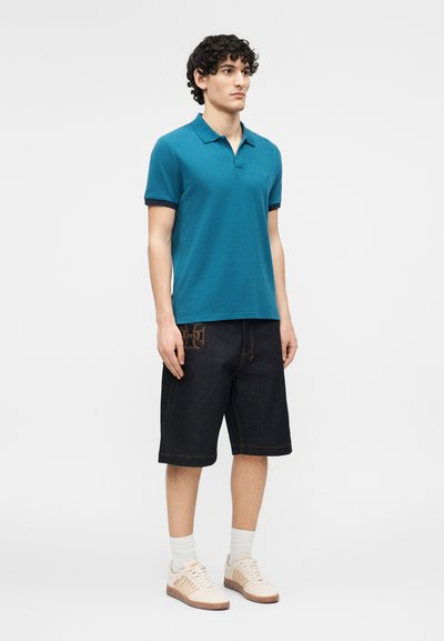 Νεαρός άντρας στέκεται, φοράει μπλε-πράσινο πόλο με κοντά μανίκια, μαύρα σορτς μέχρι το γόνατο με κέντημα, λευκές κάλτσες και μπεζ αθλητικά παπούτσια.
