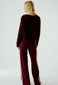 Pull en tricot bordeaux avec un motif de vagues texturé, associé à un pantalon bordeaux assorti avec des rayures blanches sur les côtés. Coupe ample, col rond.