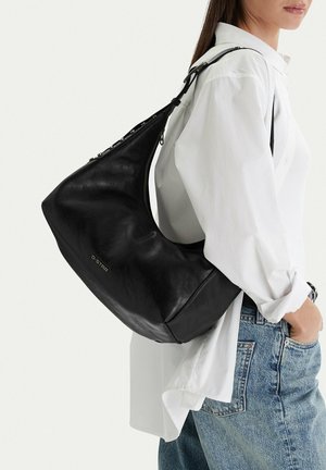 Žena v bílém tričku a modrých džínách nese na pravém rameni velkou černou koženou kabelku značky G-Star.