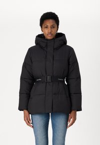 Black puffer jacket with a hood, cinched waist by a belt, featuring quilted design and logo accent on the belt. Worn with blue jeans.