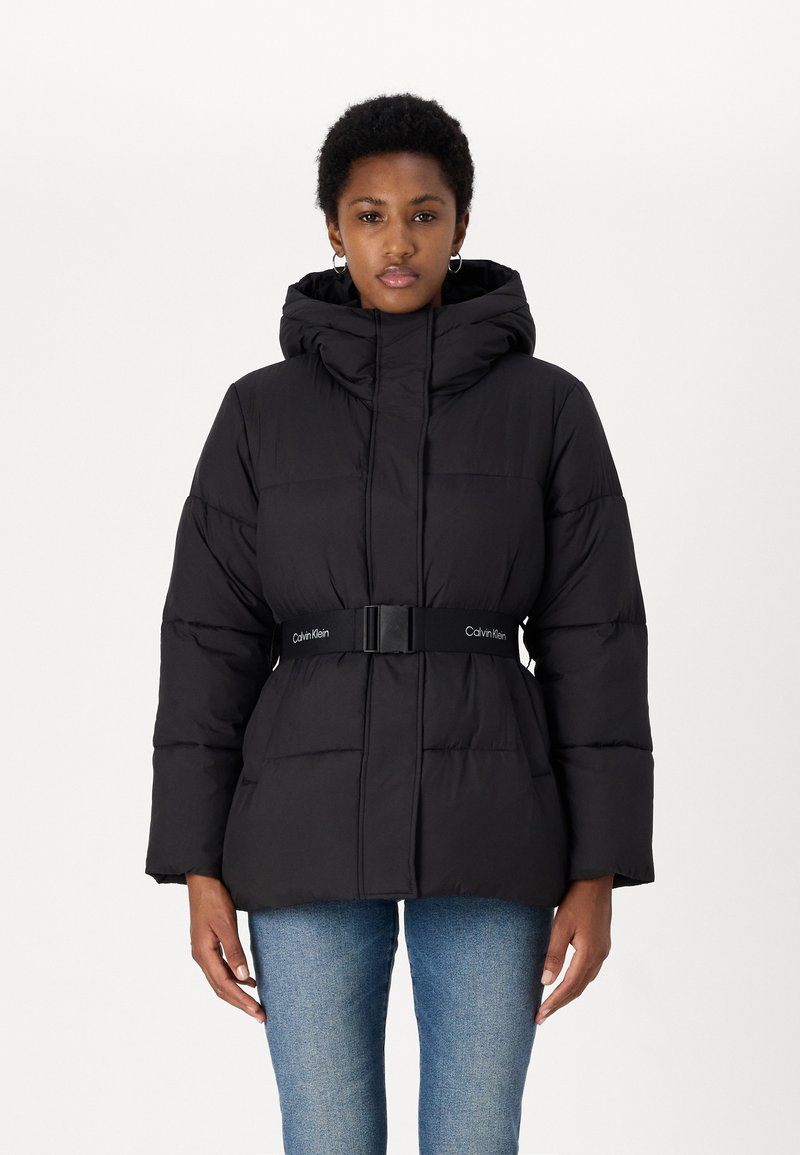Black puffer jacket with a hood, cinched waist by a belt, featuring quilted design and logo accent on the belt. Worn with blue jeans.