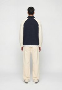 Άνδρας με την πλάτη, φοράει ένα πουλόβερ πλεκτό σε μπλε ναυτικό και εκρού, φαρδιά εκρού παντελόνια και λευκά αθλητικά παπούτσια, μπροστά σε απλό φωτεινό φόντο.