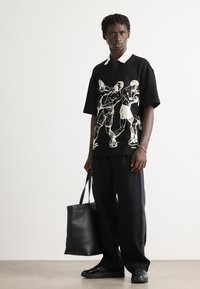 Man wearing black polo shirt with white boxing figure prints, black wide-leg pants, black shoes, holding large black leather tote bag.