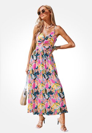 Femme portant une robe longue maxi colorée à fleurs avec fines bretelles, lunettes de soleil, collier en coquillages, sandales, et tenant un sac tissé en pose détendue.