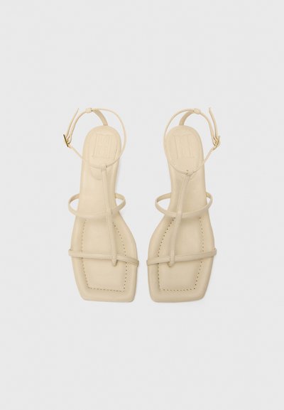 Sandalias de cuero beige con puntas cuadradas planas, tiras finas y un cinturón en el tobillo. Textura suave con detalles de costura visibles.