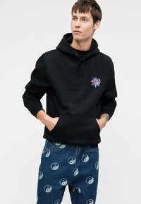 Fiatal férfi fekete Santa Cruz kapucnis pulóverben és kék farmert visel, yin-yang szimbólumokkal, kezét a pulóver zsebében tartja, és enyhén jobbra néz.