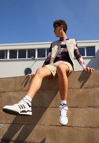 Lys beige vest over en mønstret langærmet skjorte i nuancer af lilla og rosa, parret med beige shorts og sort-hvide højhalsede sneakers.