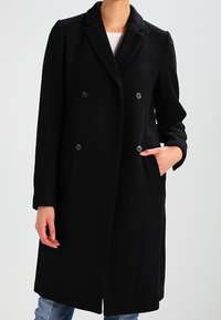 Musta villasekoitekot, joka esittelee kaksirivisen muotoilun, lovilaput, etutaskut ja nappiaksentit. Tasainen rakenne, polvipituus.