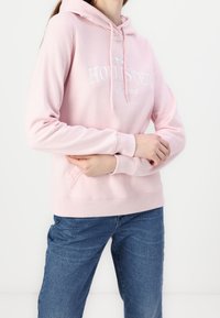 Osoba oblečená v růžové mikině Hollister s přední kapsou a modrými džíny, s překříženýma rukama na pase před bílým pozadím.