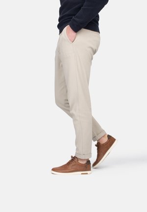 Pantalones beige ajustados con bolsillos laterales, con dobladillos rematados, combinados con zapatillas de cuero marrón con suelas blancas y diseño de cordones.